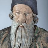 Drewniana rzeźba popiersia mężczyzny w podeszłym wieku, z długą brodą i wąsami, z czapką na głowie spod której wystają dłuższe kręte włosy. Wzrok ma spuszczony.