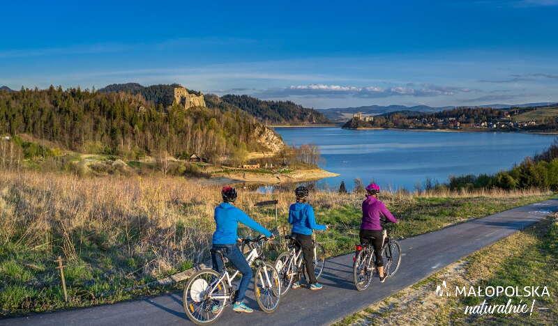 Trzech rowerzystów na ścieżce rowerowej z widokiem na Jezioro Czorsztyńskie, zamek w Czorsztynie i Niedzicy