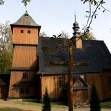 Drewniany kościół z kwadratową wieżą z przodu i dachem krytym gontem, widziany z boku, przez pryzmat stojącego obok drewnianego krzyża. Wokół rosną drzewa.
