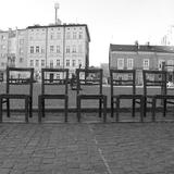 Zdjęcie czarno białe. Plac wybrukowany. Na nim stoi na wprost siedem krzeseł. Za nimi, za placem, rząd  budynków w zabudowie szeregowej.