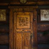 Zdobione góralską snycerką drewniane drzwi w drewnianej ścianie. Nad nimi i po bokach obrazy.