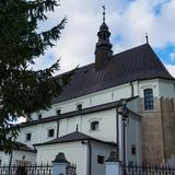 Boczna elewacja dużego wiejskiego kościoła murowanego. Białe ściany, czarne dachy. Kościół otacza metalowe ogrodzenie. Po lewej stronie widok na kościół przysłaniają gałęzie drzewa.