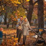Widok na mężczyznę, który w ręku trzyma liście i rzuca nimi, obok stoik kobieta uśmiecha się. Mężczyzna trzyma ją w pasie.  Obok stoi wózek dziecięcy. Z lewej stoi dziecko i bawi się liśćmi. Wokół drzewa i kolorowe liście.