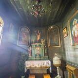 Kaplica wewnątrz drewnianego kościoła z ołtarzem w kolorze zieleni/turkusu i biało-złotą chrzcielnicą. Na drewnianych ścianach wiszą obrazy. Całość zachowana w kolorach ciemnej zieleni.