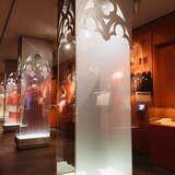 Ciemne pomieszczenie z podświetlonymi szklanymi gablotami z sutannami Ojca św. Jana Pawła II. Z tyłu widać szklane gabloty z zapisanymi  kartkami oraz duże wiszące zdjęcie księży.