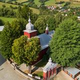 Drewniany kościół o czerwonych ścianach i jasnym dachu, stojący wśród drzew.