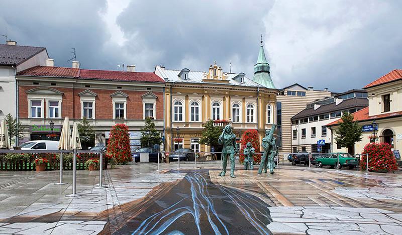 Zdjęcie przedstawia plytę Rynku Górnego w Wieliczce pokrytą trójwymiarowym malowidłem przedstawiającym wnętrze kopalni. W tle kamieniczki przy Rynku.