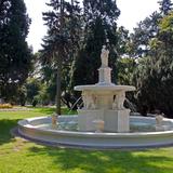 Biała fontanna z rzeźbami lwów oraz kobiety w parku. Wokół zieleń zagospodarowana.