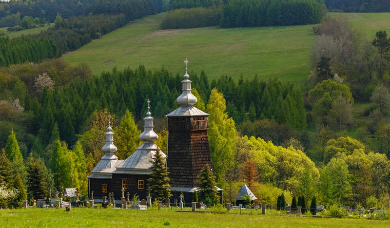 Drewniana cerkiew o ciemnych ścianach i jasnym dachu, z wieżą. Przy niej niewielki cmentarz. Wokół zielone pola i lasy.