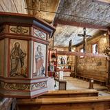 Wnętrze kościoła. Drewniana ambona z wizerunkami Ewangelistów, w tle ołtarz główny. Ściany i sufit pokryte malowidłami w odcieniach brązu.