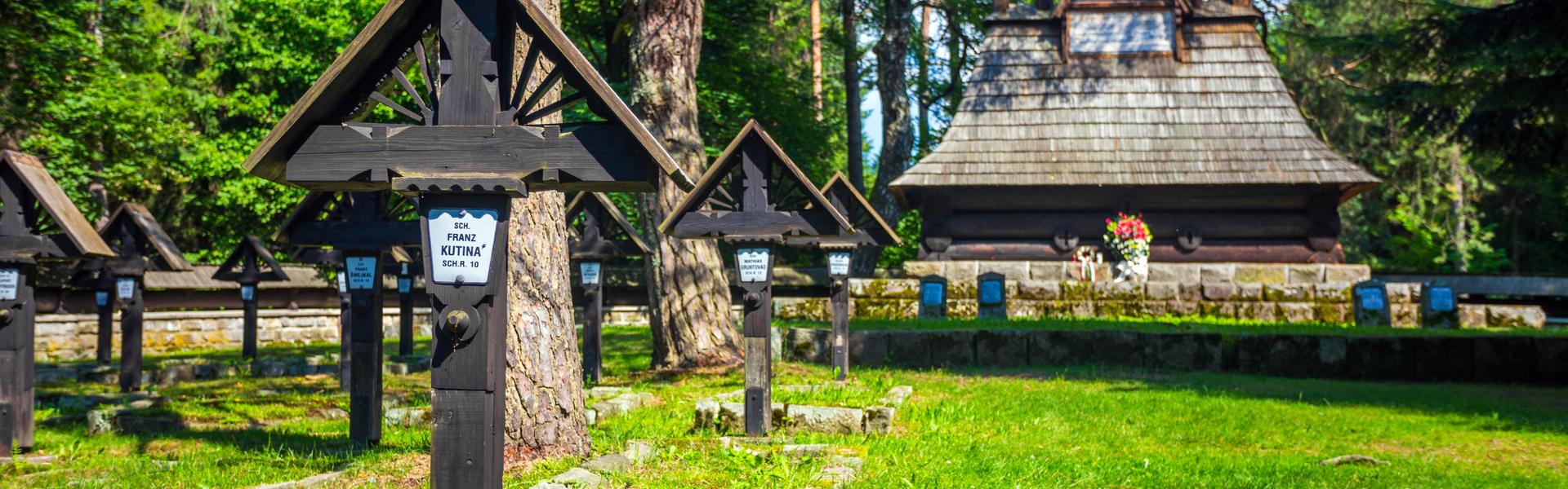 Cmentarz z wieloma drewnianymi krzyżami z białymi tabliczkami z nazwiskami, pomiędzy drzewami, po lewej, oraz po prawej z tyłu, z drewnianą kaplicą z wizerunkiem Matki Bożej w szczycie na dachu. Nad nią daszek postawiony na pionowych i poziomych belkach. Naokoło kamienne ogrodzenie. Za drzewami pomiędzy gałęziami przebija niebo.