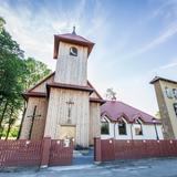 Drewniany kościół z wysoką wieżą nad wejściem. Obok murowana dzwonnica, z tyłu murowane zabudowania parafialne. Kościół otacza drewniane ogrodzenie, po lewej stronie rosną drzewa.