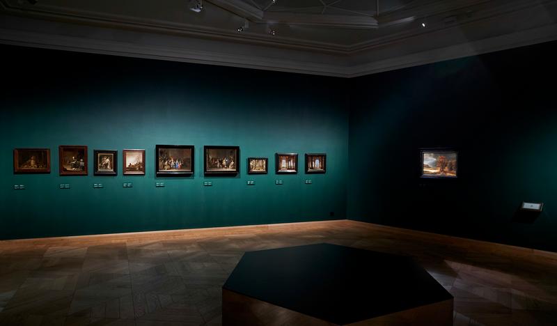 Ciemna sala wystawowa o ścianach w kolorze morskim, punktowe światło eksponuje obrazy zawieszone wzdłuż ścian. Po prawej stronie 
