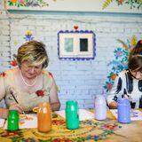 Dwie kobiety siedzące za stołem malują farbami na kartkach zalipiańskie wzory