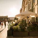 Zewnętrzny ogródek Restauracji Bianca w Krakowie z widokiem na płytę Rynku Głównego i Sukiennice. Wokół ogródka rosną rośliny w donicach, a nad stolikami znajdują się parasole z nazwą restauracji.