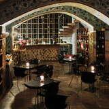 Ciemna sala w Restauracji Pod Różą w Krakowie. Na środku znajdują się czarne stoliki oraz krzesła. Na ścianach na półkach znajdują się wina. Z samego tyłu sami widać kolekcję poukładanych win.