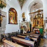 Ławki oraz krzesła w rzędach w Kościele św. Wojciecha w Krakowie. Kilka kobiet siedzi w ławkach. Na ścianach bogate, złote zdobienia oraz elementy z kamienia.