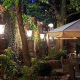 Ogródek przy restauracji Willa Fryderyka - stoliki pod parasolami, pod wiekowymi drzewami przy ceglanym murze oświetlonym stojącymi lampami