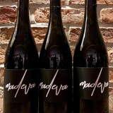Trzy stojące obok siebie butelki z Winnicy Madeva oraz etykietami z takową nazwą na tle ceglanej ściany.