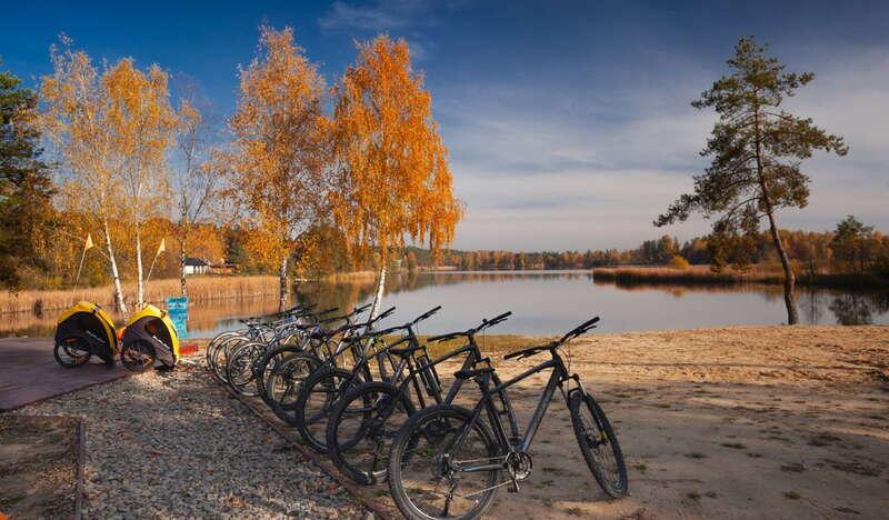 Rowery z wypożyczalni oraz przyczepki rowerowe na tle jeziora i drzew w jesiennych barwach.