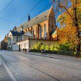 Na pierwszym planie widać ulicę z kostki brukowej wraz z torami tramwajowymi przy plantach. Zaraz za nią widać budynek Bazyliki Trójcy Świętej w Krakowie.