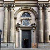 Żelazne drzwi w bramie prowadzącej do wnętrza Kościoła św. Anny w Krakowie. Obok drzwi po jednej jak i drugiej stronie znajdują się po dwie wysokie, korynckie kolumny. Nad drzwiami tablica i wyżej okno z łukiem.