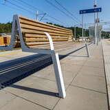 Metalowa ławka z drewnianym siedziskiem ustawiona na peronie kolejowym.