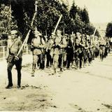 Historyczne zdjęcie w sepii - kolumna idących żołnierzy ze strzelbami. Po obu stronach drogi drzewa.