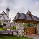 Brama i za nią kościół, kamienne z elementami drewnianymi, w stylu regionalnym góralskim. Współczesny w otoczeniu zieleni.